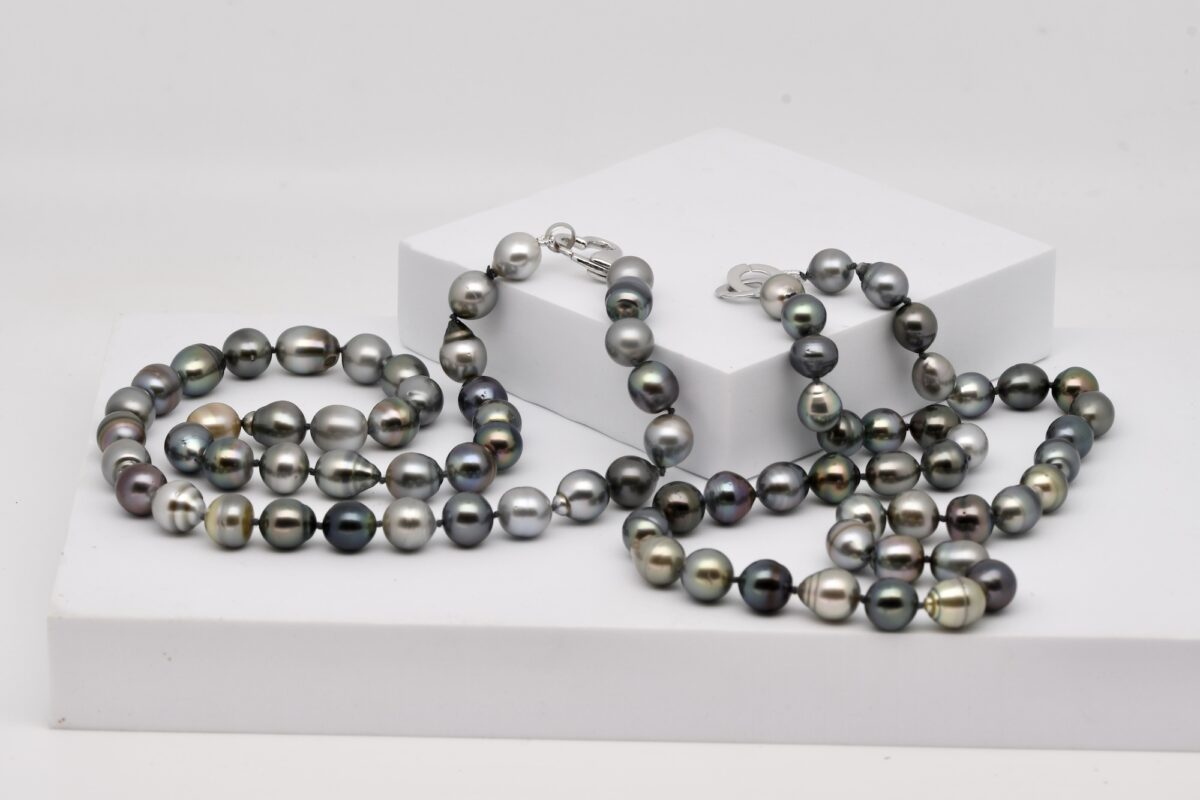 Photographié sur fond blanc, les colliers de perles Tetae et Maeaea se présentent en courbe douce, formé de perles de Tahiti semi baroques ou cerclées aux nuances naturelles variées : vert, gris, bleu, aubergine, bronze. Le montage à nœuds réguliers entre chaque perle assure un tombé souple. Les perles, de 10 à 11 mm, sont légèrement irrégulières, ce qui renforce leur caractère. À l’extrémité, un fermoir en argent 925 rhodié ajoute une touche sentimentale et brillante à l’ensemble.