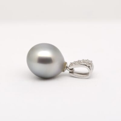 Sur fond blanc, le pendentif Alika révèle une perle de Tahiti ovale bleu nuit foncé, aux légères marques naturelles de surface, montée sur une bélière ouvragée en argent 925 rhodié. La photo met en valeur le lustre excellent de la perle, captant la lumière en reflets bleu roi et gris profond. Le fond neutre fait ressortir la pureté du métal et la profondeur des nuances marines de la perle.