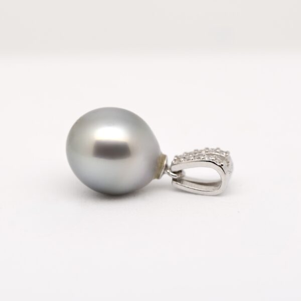 Sur fond blanc, le pendentif Alika révèle une perle de Tahiti ovale bleu nuit foncé, aux légères marques naturelles de surface, montée sur une bélière ouvragée en argent 925 rhodié. La photo met en valeur le lustre excellent de la perle, captant la lumière en reflets bleu roi et gris profond. Le fond neutre fait ressortir la pureté du métal et la profondeur des nuances marines de la perle.