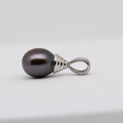 Sur fond blanc, le pendentif perle Alika révèle une perle de Tahiti ovale bleu nuit foncé, aux légères marques naturelles de surface, montée sur une bélière ouvragée en argent 925 rhodié. La photo met en valeur le lustre excellent de la perle, captant la lumière en reflets bleu roi et gris profond. Le fond neutre fait ressortir la pureté du métal et la profondeur des nuances marines de la perle.