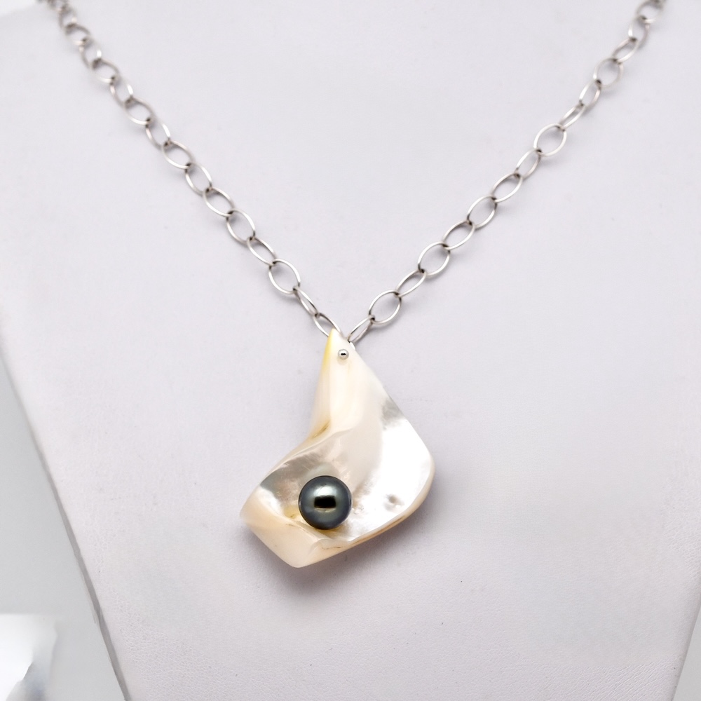 Le pendentif de ce collier est une pièce imposante de 5 × 4 cm, taillée directement dans la coquille d’une huître perlière provenant de la ferme de Manihi. La nacre, d’un blanc lumineux aux reflets irisés, a été sculptée de façon à créer un véritable écrin naturel pour la perle. Au centre du pendentif vient se loger une perle de Tahiti ronde de 9,7 mm, de qualité B, présentant de magnifiques reflets naturels vert profond.